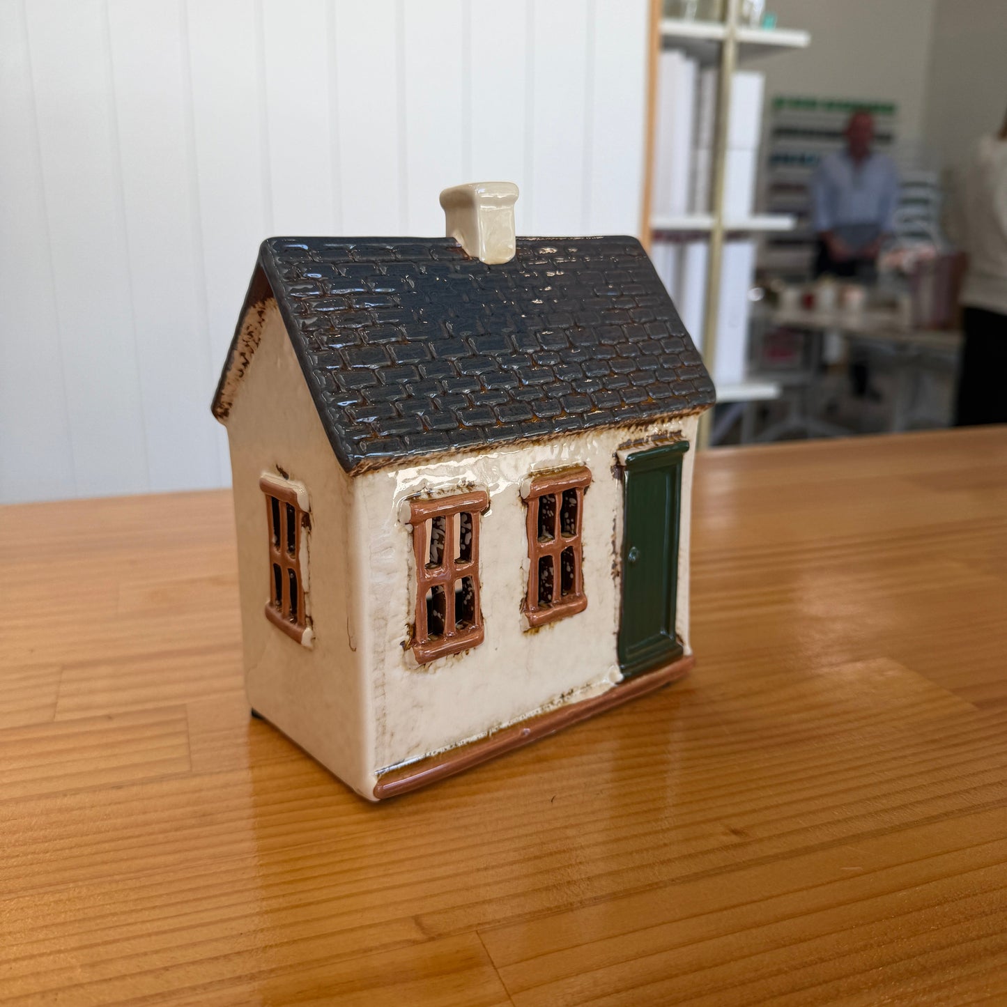 A small decorative ceramic house figurine with a dark textured roof, cream-colored walls, brown window frames, and a green door, displayed on a wooden tabletop.